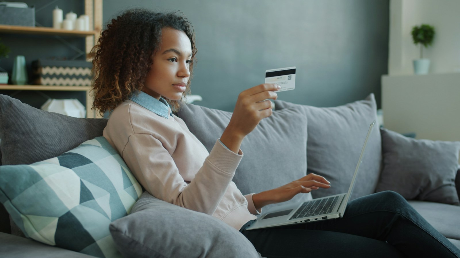 A woman on a sofa holding a credit card while shopping on her laptop