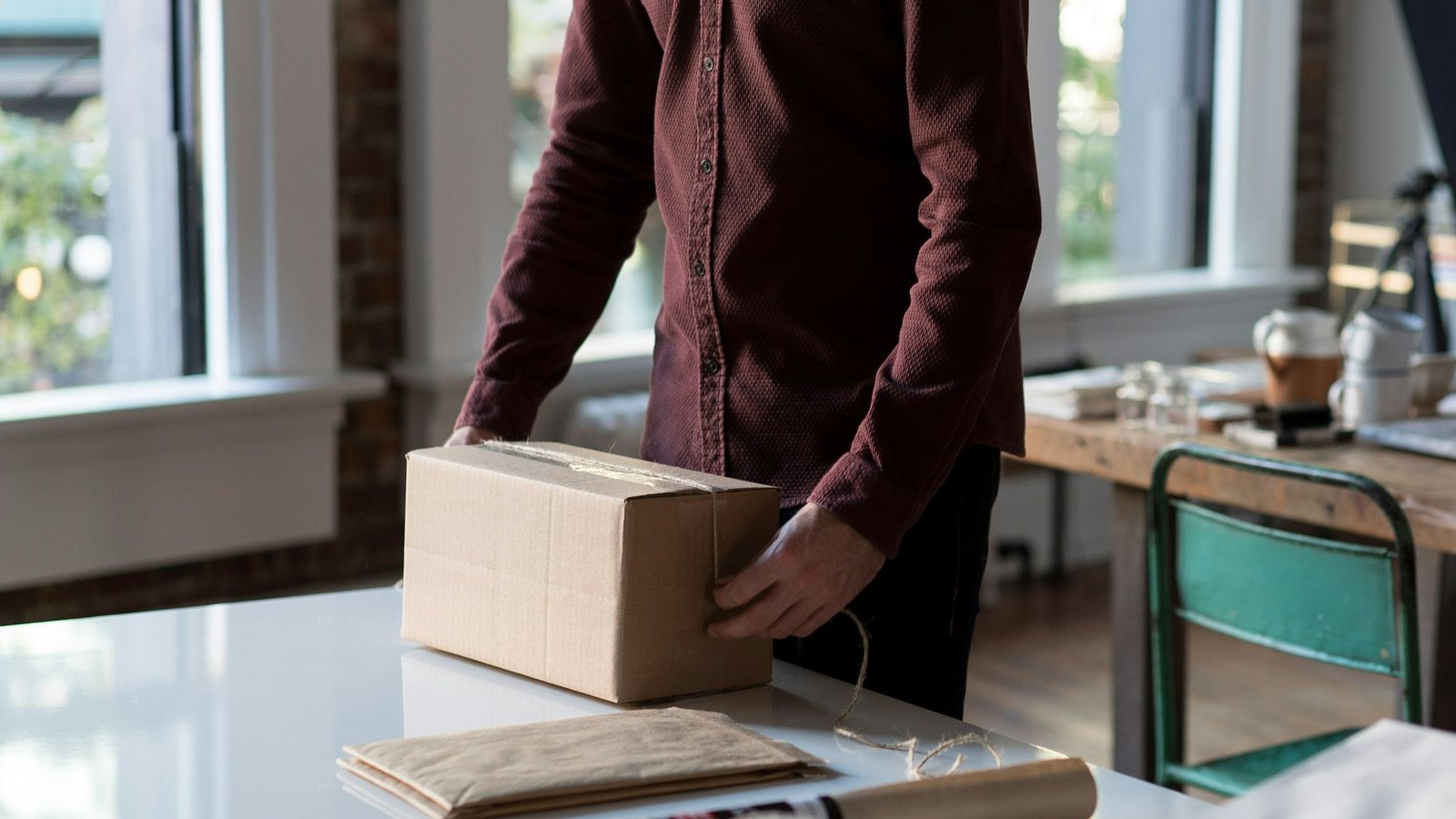 A person in a burgundy shirt tying up a brown parcel on a white table in a bright studio with tall windows