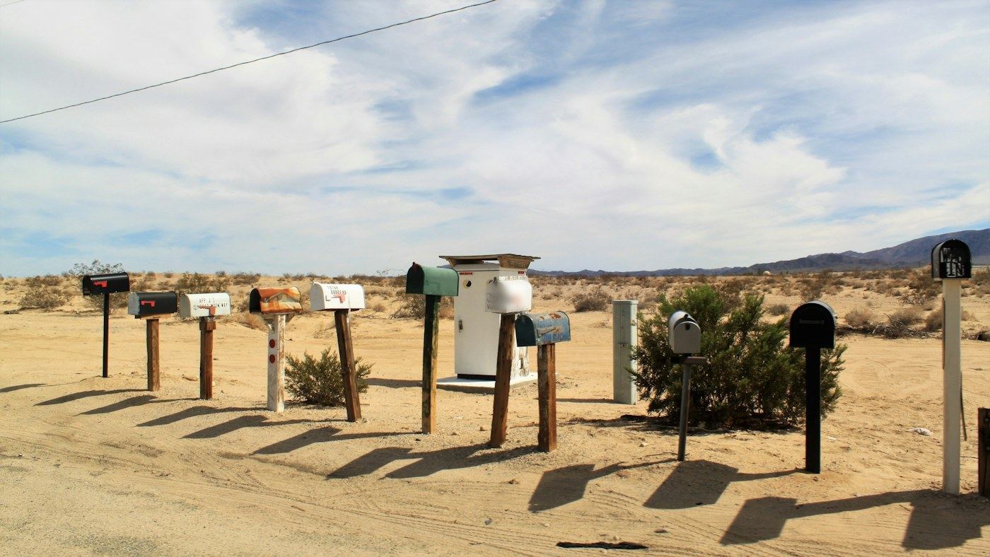 A row of mismatched roadside mailboxes on wooden posts in sandy desert under a wide sky