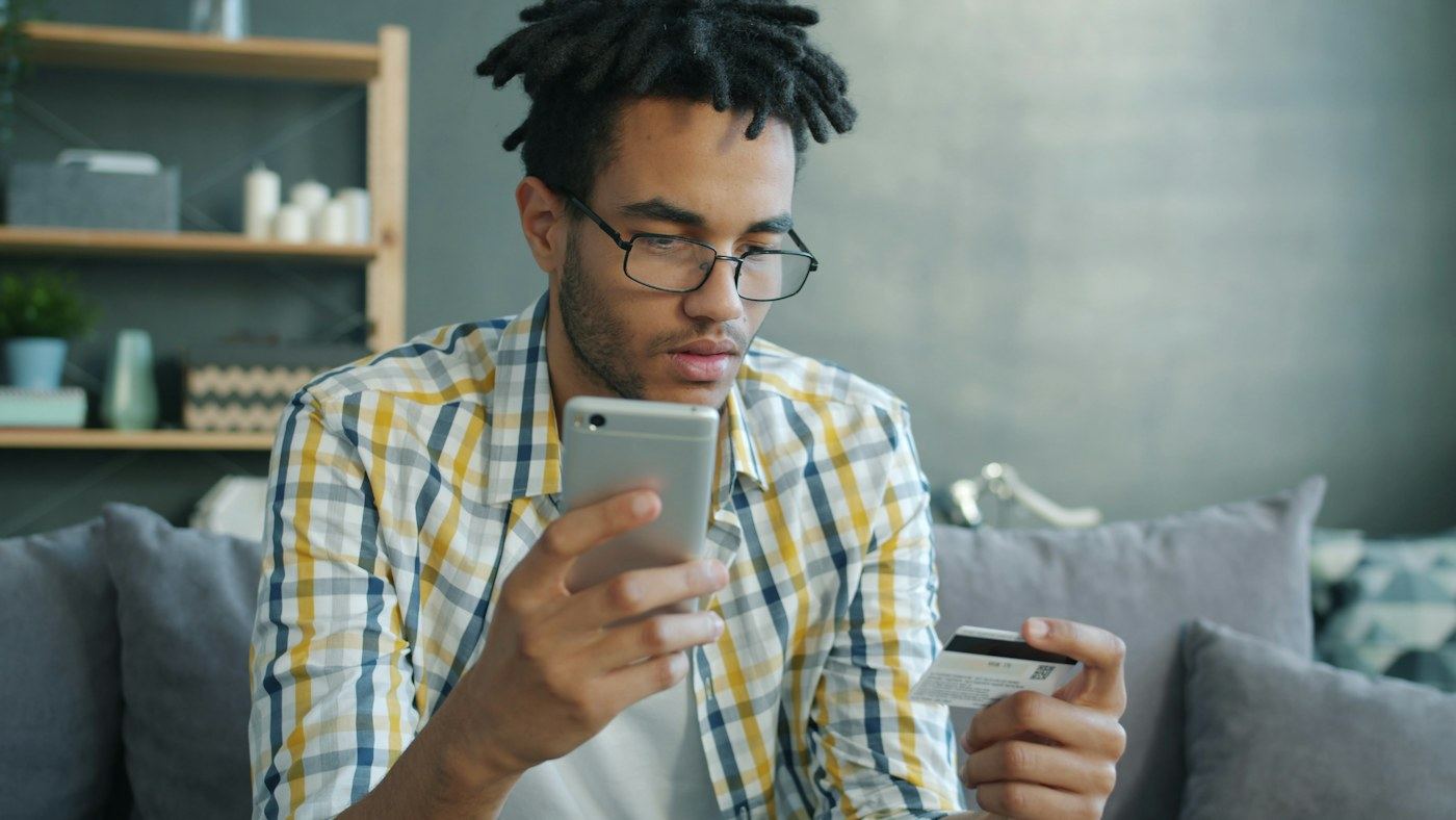 A man in glasses and a plaid shirt on a gray sofa, holding a phone in one hand and a credit card in the other